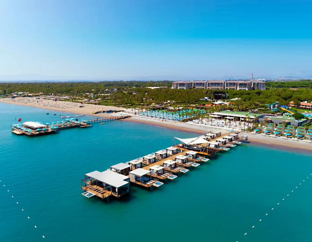 Luftaufnahme des Regnum The Crown Belek mit Privatstrand, luxuriösen Badepavillons auf dem Wasser und Blick auf das weitläufige Resort